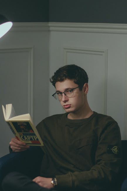 A man wearing glasses sitting in a chair reading a book.